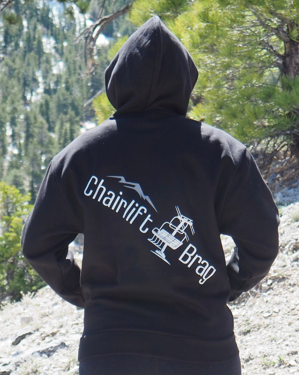 Person wearing a black hoodie with 'Chairlift Brag' text and logo in a forest setting