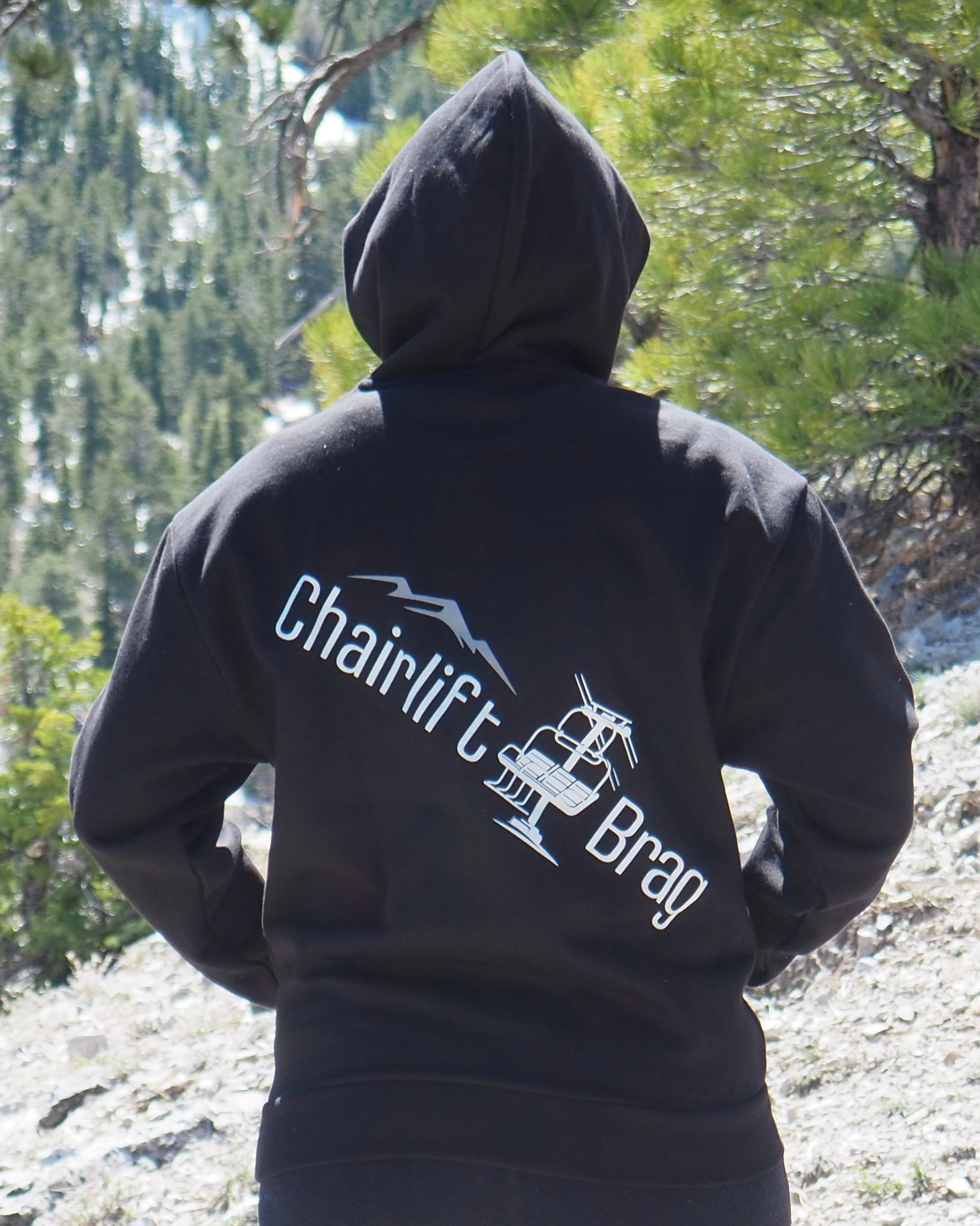 Person wearing a black hoodie with 'Chairlift Brag' text and logo in a forest setting