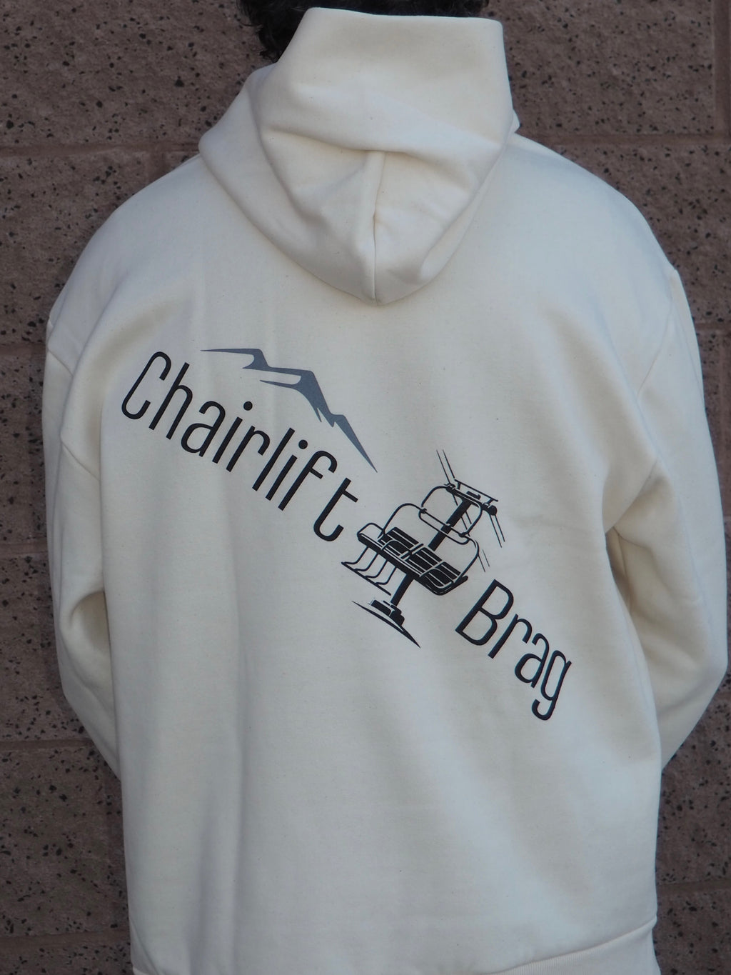 White hoodie with 'Chairlift Brag' graphic on back in front of  a textured brown background