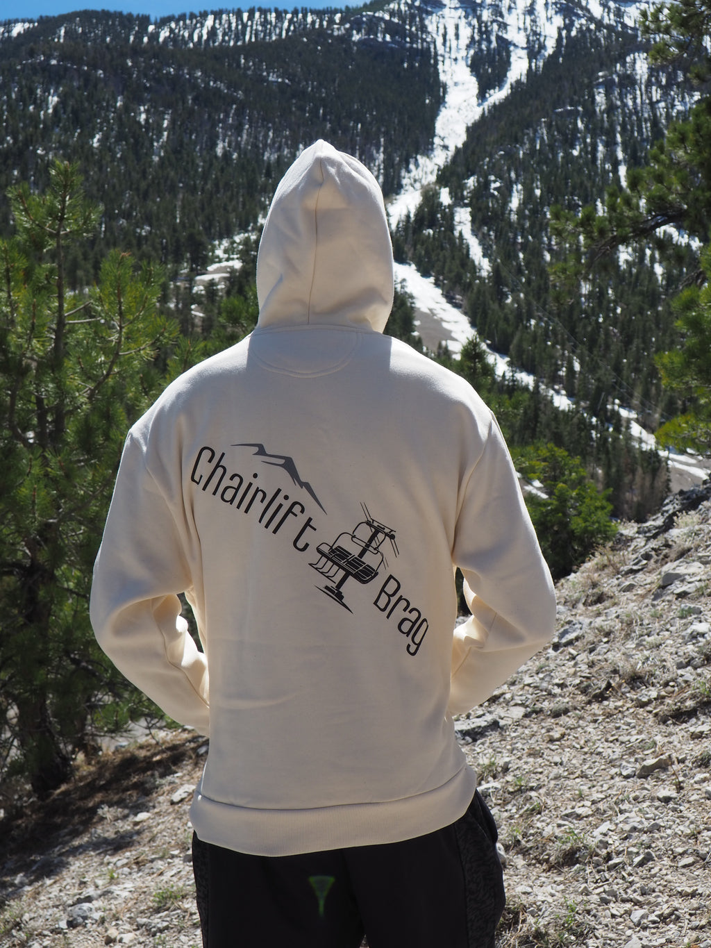 Person wearing a off white hoodie with Chairlift Brag text and graphics in a mountainous area