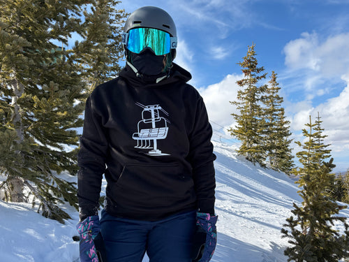 Person in ski gear with a black Chairlift Brag hoodie featuring a chairlift design, standing in a snowy landscape with trees.