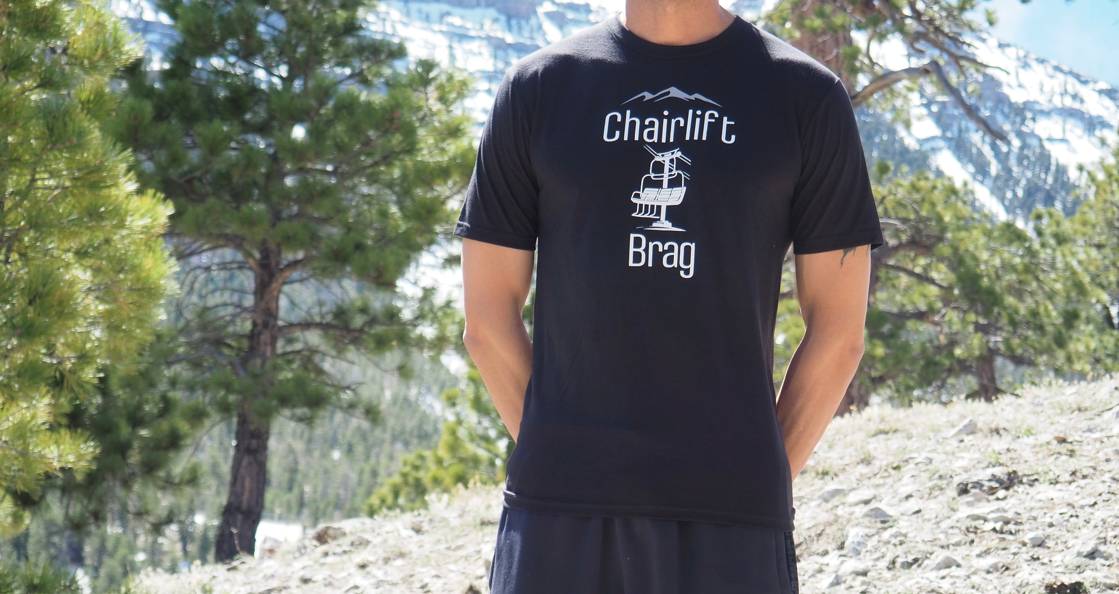 Person wearing a black t-shirt with 'Chairlift Brag' text and graphic, standing outdoors with trees and mountains in the background.
