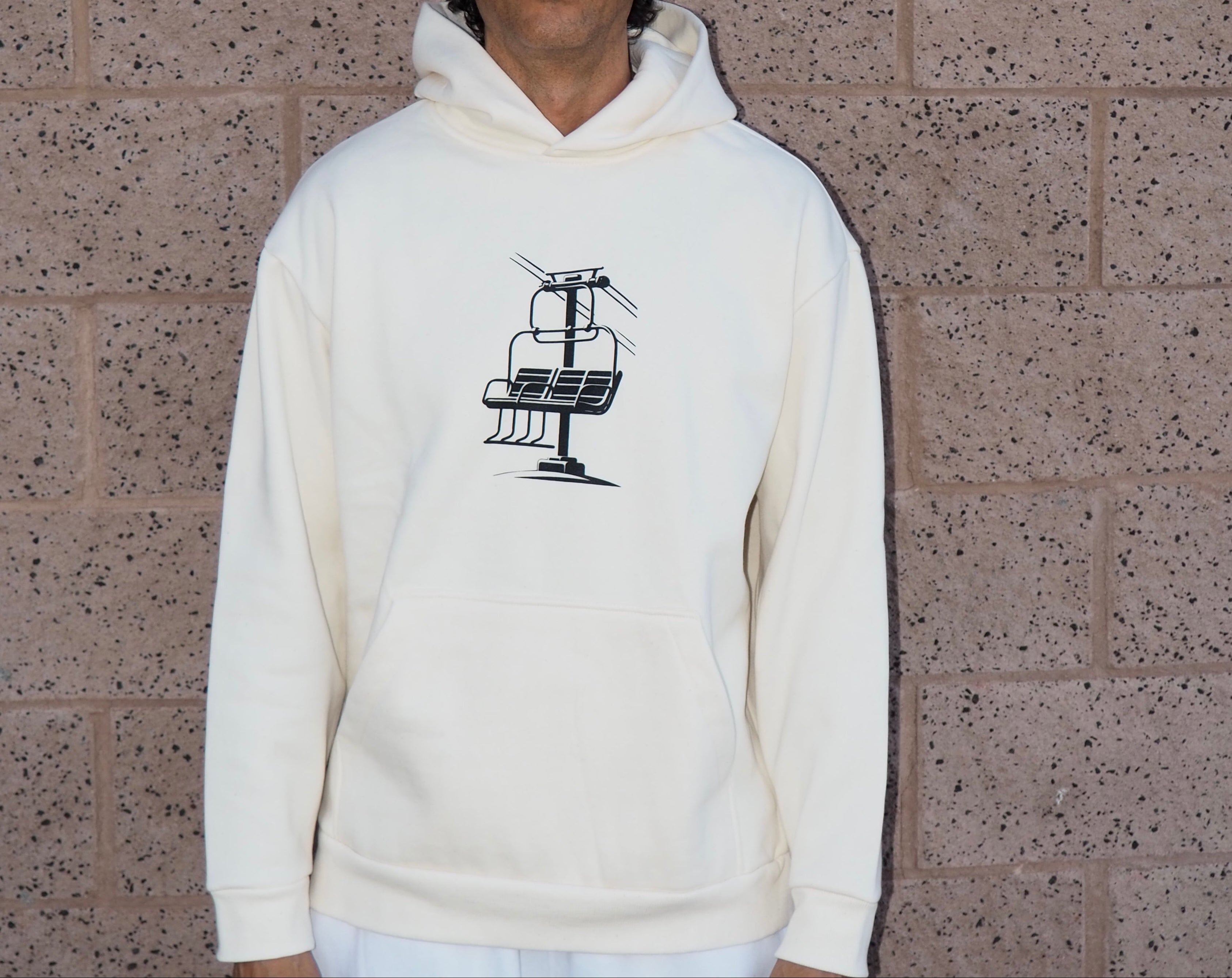 Person wearing a white hoodie with a Chairlift Brag graphic design against a textured wall.