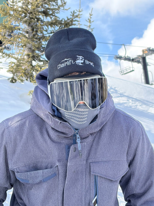 Person wearing a gray jacket, black Chairlift Brag beanie, and goggles with a snowy landscape in the background.