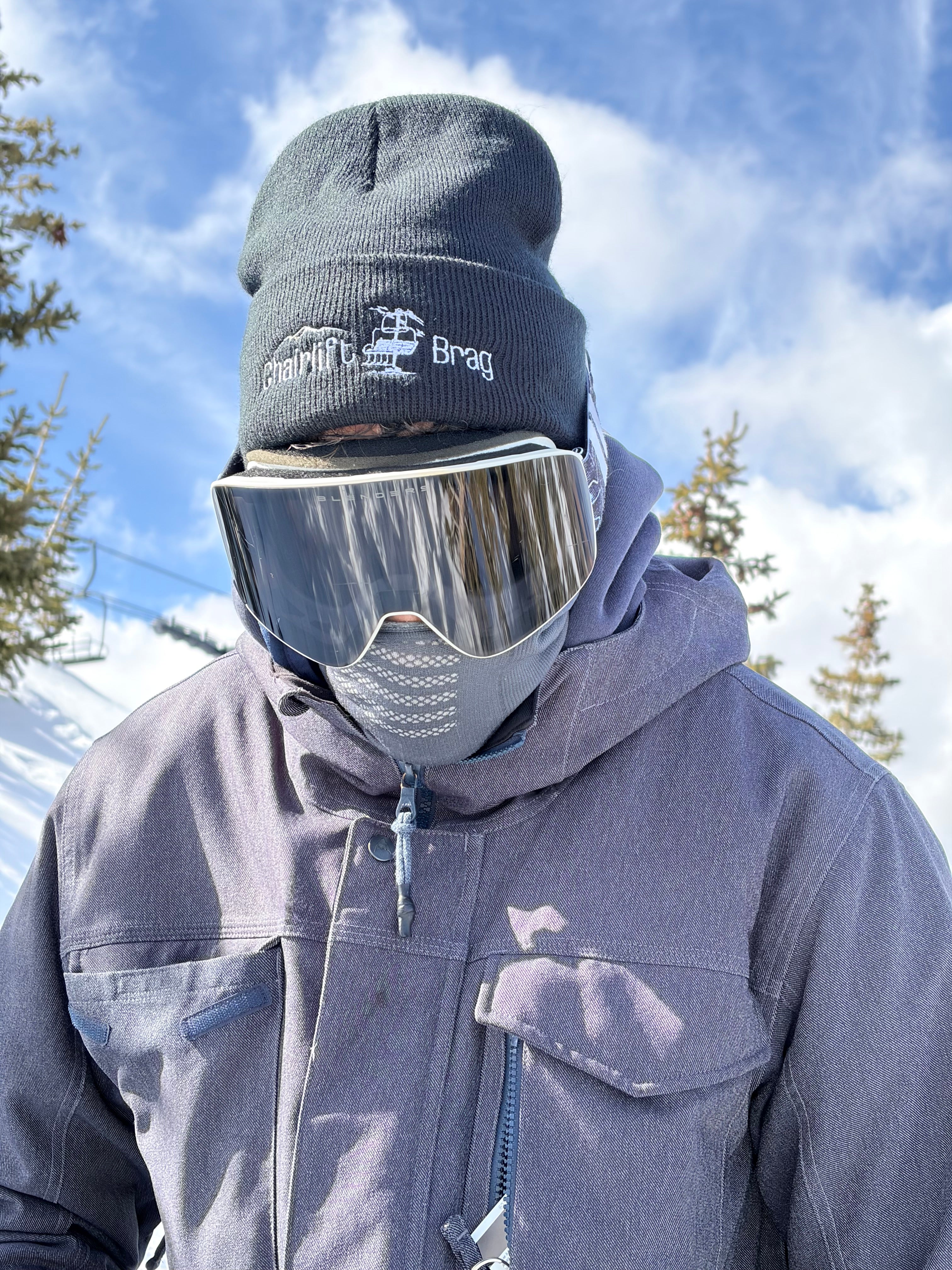 Person wearing a gray jacket and  Chairlift Brag beanie and goggles with a blue sky and trees in the background